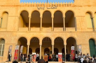 ​لقطة جماعية لافتتاح المعرض الوطني للفنون التشكيلية "رؤى معاصرة" أمام واجهة مبنى قصر الخلد الأثري في طرابلس، يظهر فيها الحضور والمنظمون على منصة أمام الأقواس التاريخية للمبنى مع وجود كاميرات تصوير وتغطية إعلامية وشعار منصة الصباح
