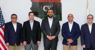خمسة مسؤولين ورجال أعمال/عسكريين يرتدون بدلات رسمية يقفون جنبًا إلى جنب أمام خريطة ليبيا وعلمي ليبيا والولايات المتحدة الأمريكية.