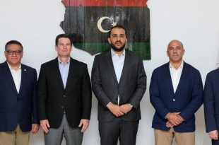 خمسة مسؤولين ورجال أعمال/عسكريين يرتدون بدلات رسمية يقفون جنبًا إلى جنب أمام خريطة ليبيا وعلمي ليبيا والولايات المتحدة الأمريكية.