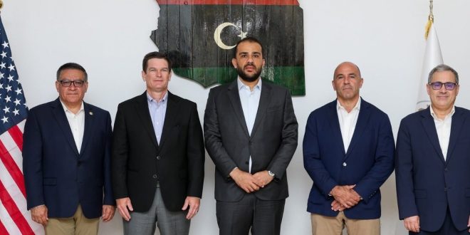 خمسة مسؤولين ورجال أعمال/عسكريين يرتدون بدلات رسمية يقفون جنبًا إلى جنب أمام خريطة ليبيا وعلمي ليبيا والولايات المتحدة الأمريكية.