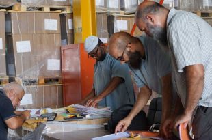 ثلاثة رجال يفحصون أوراقاً ومستندات في مخزن مليء بصناديق الكتب المدرسية لإتمام عملية التوزيع.