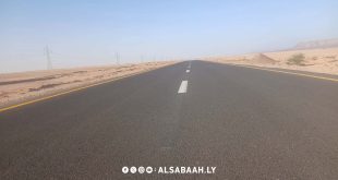 لقطة واسعة ممتدة تظهر مسار طريق إسفلتي حديث ممتد وسط بيئة صحراوية في الجنوب الليبي،