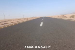 لقطة واسعة ممتدة تظهر مسار طريق إسفلتي حديث ممتد وسط بيئة صحراوية في الجنوب الليبي،