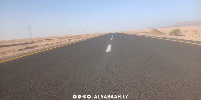 لقطة واسعة ممتدة تظهر مسار طريق إسفلتي حديث ممتد وسط بيئة صحراوية في الجنوب الليبي،