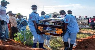 عاملون يرتدون معدات وقاية وكمامات يحملون نعشًا خلال مراسم دفن، فيما يقف عدد من الأهالي لمتابعة المشهد في منطقة مفتوحة.