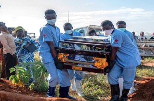 عاملون يرتدون معدات وقاية وكمامات يحملون نعشًا خلال مراسم دفن، فيما يقف عدد من الأهالي لمتابعة المشهد في منطقة مفتوحة.