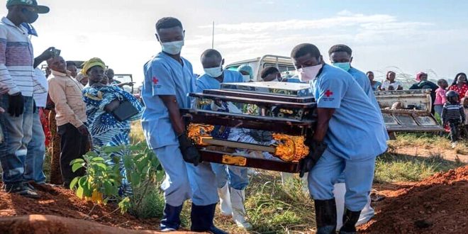 عاملون يرتدون معدات وقاية وكمامات يحملون نعشًا خلال مراسم دفن، فيما يقف عدد من الأهالي لمتابعة المشهد في منطقة مفتوحة.