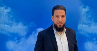 رجل يرتدي بدلة كحلية وقميصًا أبيض، ينظر إلى الكاميرا، يقف أمام خلفية زرقاء بتصميم تجريدي.
