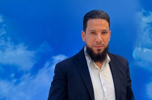 رجل يرتدي بدلة كحلية وقميصًا أبيض، ينظر إلى الكاميرا، يقف أمام خلفية زرقاء بتصميم تجريدي.