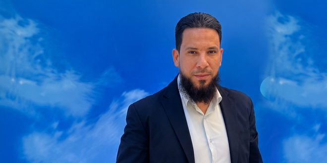 رجل يرتدي بدلة كحلية وقميصًا أبيض، ينظر إلى الكاميرا، يقف أمام خلفية زرقاء بتصميم تجريدي.