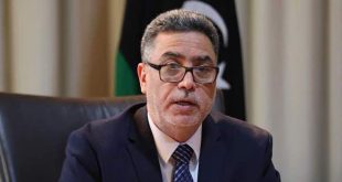 صورة الخبير الاقتصادي رجب خليل يرتدي نظارة وبدلة رسمية، وأمامه العلم الليبي في الخلفية