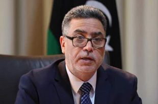 صورة الخبير الاقتصادي رجب خليل يرتدي نظارة وبدلة رسمية، وأمامه العلم الليبي في الخلفية