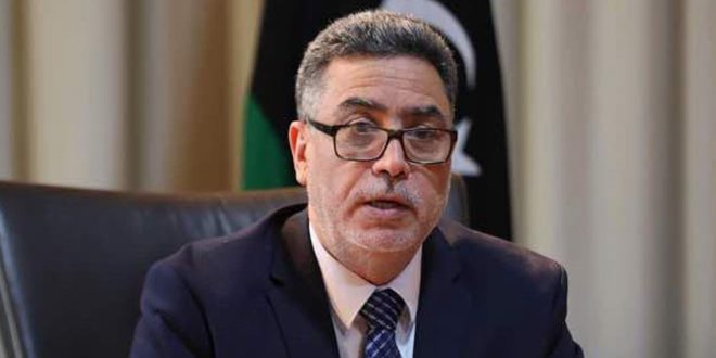 صورة الخبير الاقتصادي رجب خليل يرتدي نظارة وبدلة رسمية، وأمامه العلم الليبي في الخلفية