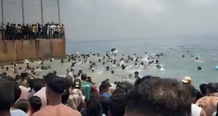 أعداد كبيرة من المهاجرين يتجمعون على شاطئ البحر وعند حاجز حديدي حدودي، بينما يسبح آخرون في مياه البحر محاولين العبور نحو الجانب الآخر