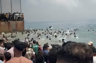 أعداد كبيرة من المهاجرين يتجمعون على شاطئ البحر وعند حاجز حديدي حدودي، بينما يسبح آخرون في مياه البحر محاولين العبور نحو الجانب الآخر