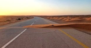 طريق زفتي في منطقة صحراوية تغطيه أجزاء من الكثبان والرمال المتحركة عند غروب الشمس.