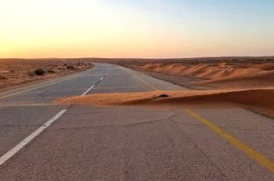 طريق زفتي في منطقة صحراوية تغطيه أجزاء من الكثبان والرمال المتحركة عند غروب الشمس.