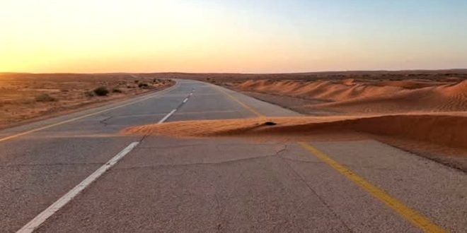 طريق زفتي في منطقة صحراوية تغطيه أجزاء من الكثبان والرمال المتحركة عند غروب الشمس.