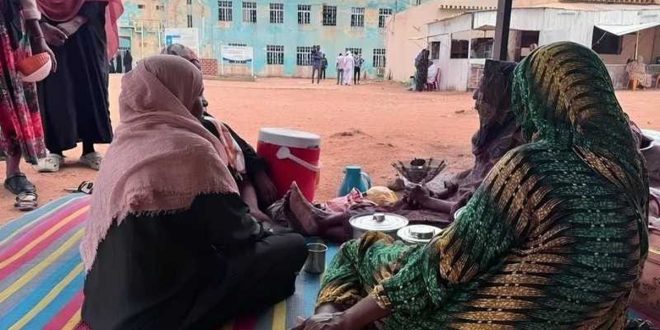 ​"نساء نازحات جالسات على حصير في ساحة عامة بمدينة الأبيض السودانية وسط ظروف إنسانية صعبة وأزمة نزوح متفاقمة."