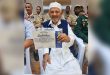 "القائد الكشفي المخضرم مسعود الهباط يمسك بشهادة تقدير وتكريم، وأمامه عدد من أعضاء وقادة الحركة العامة للكشافة والمرشدات في ليبيا بزيّهم الكشفي."