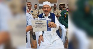 ​"القائد الكشفي المخضرم مسعود الهباط يمسك بشهادة تقدير وتكريم، وأمامه عدد من أعضاء وقادة الحركة العامة للكشافة والمرشدات في ليبيا بزيّهم الكشفي."