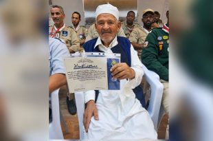 ​"القائد الكشفي المخضرم مسعود الهباط يمسك بشهادة تقدير وتكريم، وأمامه عدد من أعضاء وقادة الحركة العامة للكشافة والمرشدات في ليبيا بزيّهم الكشفي."