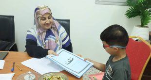 وظفة إدارية محجبة تبتسم وتسلم طفلاً صغيراً يرتدي نظارة ملف تسجيل باللون الأزرق مكتوب عليه "ملف" باللغة العربية، وذلك ضمن إجراءات التسجيل للصف الأول للعام الدراسي 2026-2027.