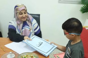 وظفة إدارية محجبة تبتسم وتسلم طفلاً صغيراً يرتدي نظارة ملف تسجيل باللون الأزرق مكتوب عليه "ملف" باللغة العربية، وذلك ضمن إجراءات التسجيل للصف الأول للعام الدراسي 2026-2027.