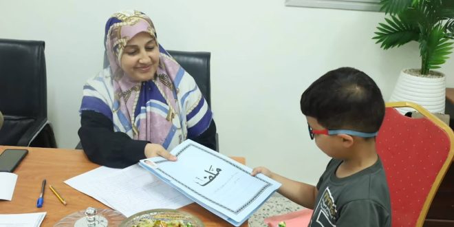 وظفة إدارية محجبة تبتسم وتسلم طفلاً صغيراً يرتدي نظارة ملف تسجيل باللون الأزرق مكتوب عليه "ملف" باللغة العربية، وذلك ضمن إجراءات التسجيل للصف الأول للعام الدراسي 2026-2027.
