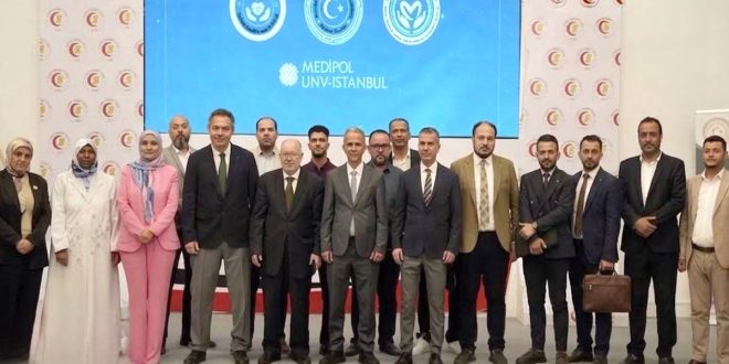 ​صورة جماعية للمشاركين والفرق الطبية الليبية والتركية في الملتقى الليبي التركي لزراعة الكبد بطرابلس أمام شاشة عرض تحمل شعار مستشفى ميديبول التركي.