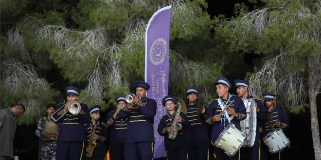 صورة خارجية لفرقة موسيقية شبابية تعزف ليلاً في حفل اختتام المخيمات الوطنية للشباب بغابات ليبيا، مع لافتة للهيئة في الخلفية.