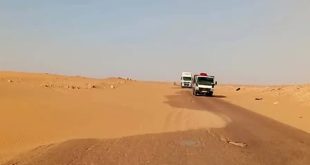 شاحنتان تسيران على طريق إسفلتي صحراوي تتراكم على أحد جوانبه الكثبان الرملية الزاحفة بين أجدابيا وطبرق.