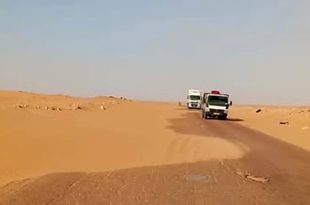 شاحنتان تسيران على طريق إسفلتي صحراوي تتراكم على أحد جوانبه الكثبان الرملية الزاحفة بين أجدابيا وطبرق.