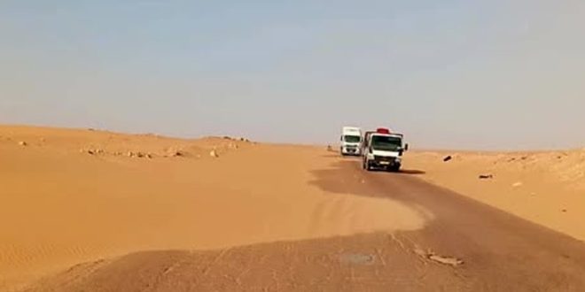 شاحنتان تسيران على طريق إسفلتي صحراوي تتراكم على أحد جوانبه الكثبان الرملية الزاحفة بين أجدابيا وطبرق.