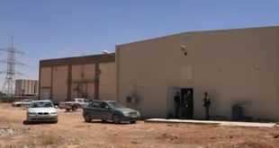 مبنى خدمي ومحطة كهرباء في حي الحدائق بمدينة طبرق، مع وجود سيارات ورقعة ترابية وبرج لنقل الكهرباء في الخلفية.