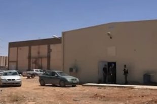 مبنى خدمي ومحطة كهرباء في حي الحدائق بمدينة طبرق، مع وجود سيارات ورقعة ترابية وبرج لنقل الكهرباء في الخلفية.