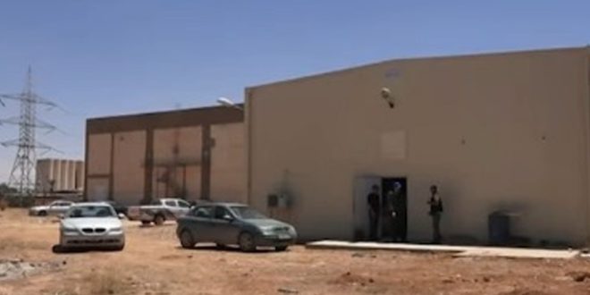 مبنى خدمي ومحطة كهرباء في حي الحدائق بمدينة طبرق، مع وجود سيارات ورقعة ترابية وبرج لنقل الكهرباء في الخلفية.