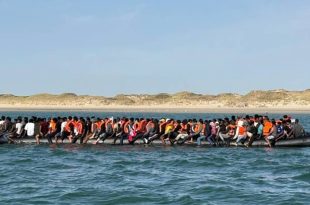 صورة توثق حادثة مأساوية لمهاجرين في البحر المتوسط، حيث يظهر زورق مطاطي أسود محمل بأعداد كبيرة من الأشخاص الذين يرتدون سترات نجاة برتقالية قبالة سواحل رملية. هذه الصورة مرتبطة بخبر العثور على 11 جثة متحللة قبالة زوارة.