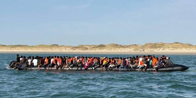 صورة توثق حادثة مأساوية لمهاجرين في البحر المتوسط، حيث يظهر زورق مطاطي أسود محمل بأعداد كبيرة من الأشخاص الذين يرتدون سترات نجاة برتقالية قبالة سواحل رملية. هذه الصورة مرتبطة بخبر العثور على 11 جثة متحللة قبالة زوارة.