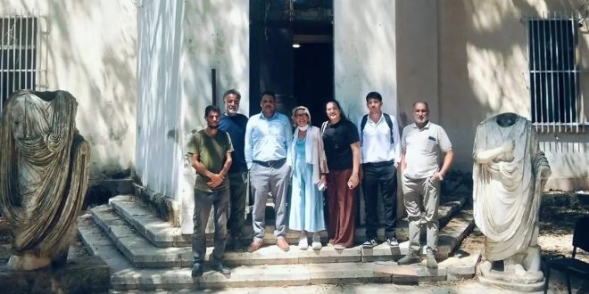 صورة جماعية لسبعة أشخاص، من بينهم خبراء من البعثة الإيطالية ومسؤولون من مراقبة آثار شحات، يقفون بابتسامة أمام مدخل مبنى أثري قديم، محاطين بتمثالين حجريين أثريين منقوشين على الجانبين. الأفراد ينظرون نحو الكاميرا، والإضاءة طبيعية.