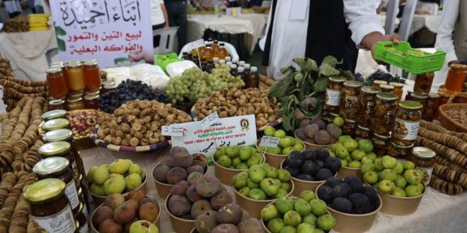 صورة متوسطة الحجم تظهر طاولة عرض غنية ومزدحمة في مهرجان "القلعة" للتين والفاكهة البعلية في جبل نفوسة، مليئة بسلال وأكواب من التين الطازج والتمور والمنتجات المحلية الأخرى مثل المربى في مرطبانات زجاجية، مع لافتات واضحة وأشخاص يتفاعلون في الخلفية.