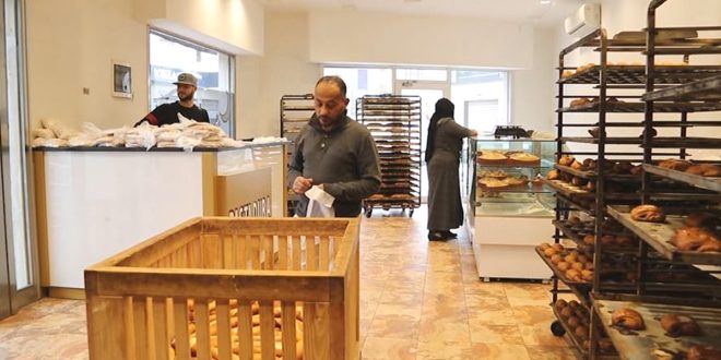 ​صورة داخلية لمخبز في ليبيا تظهر فيه أرفف المعجنات وصندوق خشبي للخبز مع وجود عمال وزبائن، تعبيرًا عن أزمة ارتفاع تكاليف الإنتاج وتراجع قيمة الدينار التي تهدد قطاع المخابز.