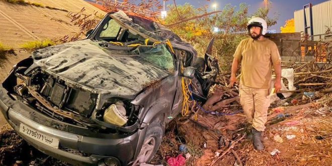 مركبة سوداء تعرضت لأضرار بالغة وتحطم في سقفها وواجهتها جراء حادث سير، وبجوارها رجل أمن يقف بموقع الحادث قرب جسر حي دمشق بطرابلس.