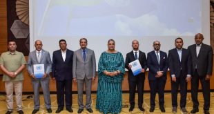 "صورة جماعية رسمية لمجموعة من ثمانية مسؤولين ليبيين، بينهم سيدتان، يبتسمون للكاميرا بعد توقيعهم على اتفاق بشأن الانتخابات الليبية. ويظهر المسؤولون واقفين أمام شعار الأمم المتحدة على شاشة خلفية. اثنان من الممثلين يرفعان وثيقة الاتفاق. المشهد يجسد أجواء من التفاؤل والإنجاز".