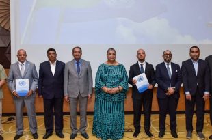 "صورة جماعية رسمية لمجموعة من ثمانية مسؤولين ليبيين، بينهم سيدتان، يبتسمون للكاميرا بعد توقيعهم على اتفاق بشأن الانتخابات الليبية. ويظهر المسؤولون واقفين أمام شعار الأمم المتحدة على شاشة خلفية. اثنان من الممثلين يرفعان وثيقة الاتفاق. المشهد يجسد أجواء من التفاؤل والإنجاز".