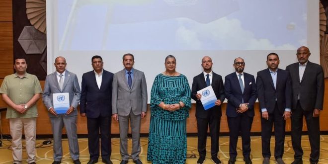 "صورة جماعية رسمية لمجموعة من ثمانية مسؤولين ليبيين، بينهم سيدتان، يبتسمون للكاميرا بعد توقيعهم على اتفاق بشأن الانتخابات الليبية. ويظهر المسؤولون واقفين أمام شعار الأمم المتحدة على شاشة خلفية. اثنان من الممثلين يرفعان وثيقة الاتفاق. المشهد يجسد أجواء من التفاؤل والإنجاز".