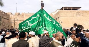 مشهد حيوي ومزدحم من احتفالات المولد النبوي في ليبيا، حيث يسير موكب كبير في ساحة عامة محاطة بأسوار تاريخية. يرفع المشاركون، ومعظمهم يرتدون الملابس التقليدية، علماً أخضر ضخماً منقوشاً بالعبارات الإسلامية. يظهر في الصورة حشود من الرجال والأطفال وهم يحتفلون بهذه المناسبة الدينية والاجتماعية.