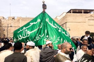 مشهد حيوي ومزدحم من احتفالات المولد النبوي في ليبيا، حيث يسير موكب كبير في ساحة عامة محاطة بأسوار تاريخية. يرفع المشاركون، ومعظمهم يرتدون الملابس التقليدية، علماً أخضر ضخماً منقوشاً بالعبارات الإسلامية. يظهر في الصورة حشود من الرجال والأطفال وهم يحتفلون بهذه المناسبة الدينية والاجتماعية.