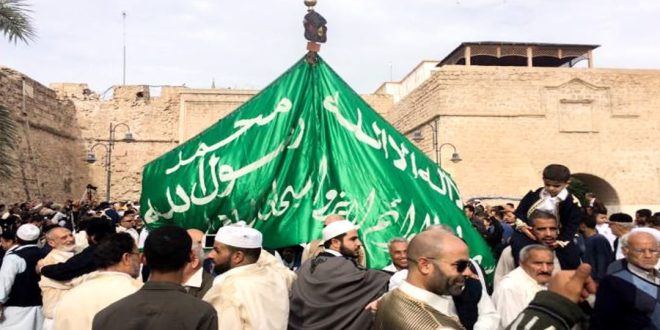 مشهد حيوي ومزدحم من احتفالات المولد النبوي في ليبيا، حيث يسير موكب كبير في ساحة عامة محاطة بأسوار تاريخية. يرفع المشاركون، ومعظمهم يرتدون الملابس التقليدية، علماً أخضر ضخماً منقوشاً بالعبارات الإسلامية. يظهر في الصورة حشود من الرجال والأطفال وهم يحتفلون بهذه المناسبة الدينية والاجتماعية.