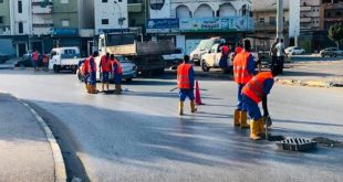 مجموعة من عمال الصيانة يرتدون سترات برتقالية وأحذية واقية أثناء صيانة وتنظيف فتحات تصريف مياه الأمطار في أحد الشوارع العامة.